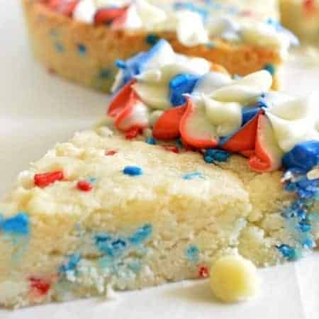 Celebrate the Fourth of July with this Patriotic Sugar Cookie Cake. This sugar cookie cake is loaded with white chocolate chips and red white and blue sprinkles! Topped with a tri-color swirled buttercream frosting!