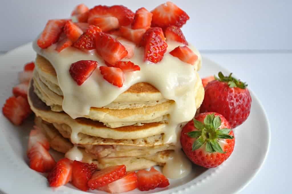 Stack of pancakes with cream cheese glaze and diced strawberries on a white plate.