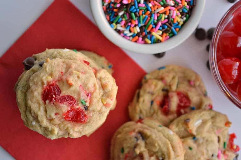 Overhead of banana split cookies with bowls of sprinkles and cherries on the side.