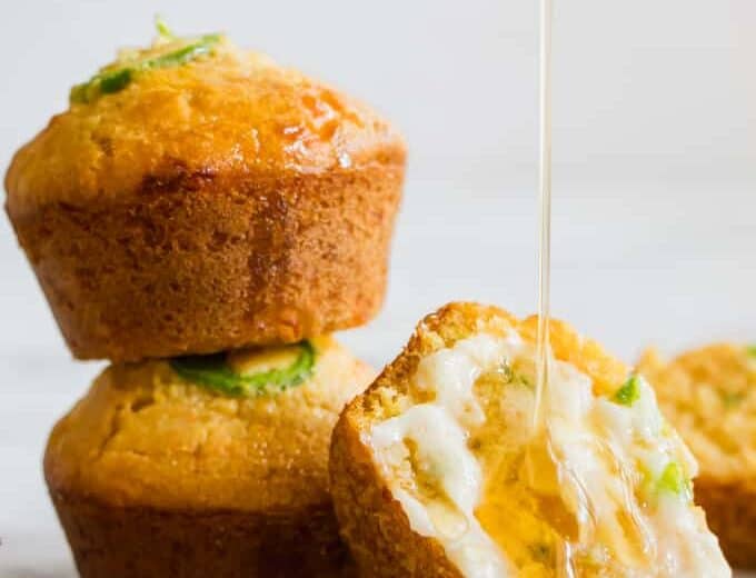 Stacked Jalapeno Cheddar Cornbread muffins, with one cut open and smeared with a sweet honey butter and a honey drizzle running down.