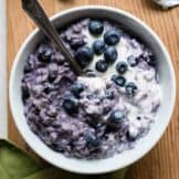 Bowl filled with purple colored blueberry oatmeal, topped with cream and fresh blueberries.