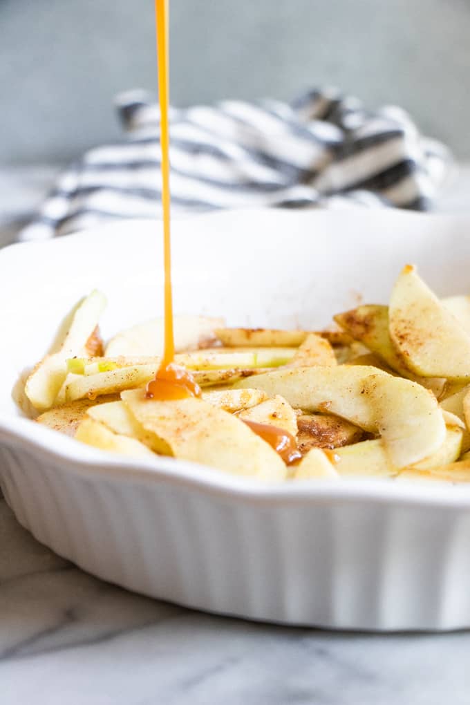 A pie dish filled with apples being drizzled with caramel for a caramel apple crisp.