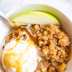 A bowl filled with caramel apple crisp, topped with ice cream drizzled with caramel and slices of apple.