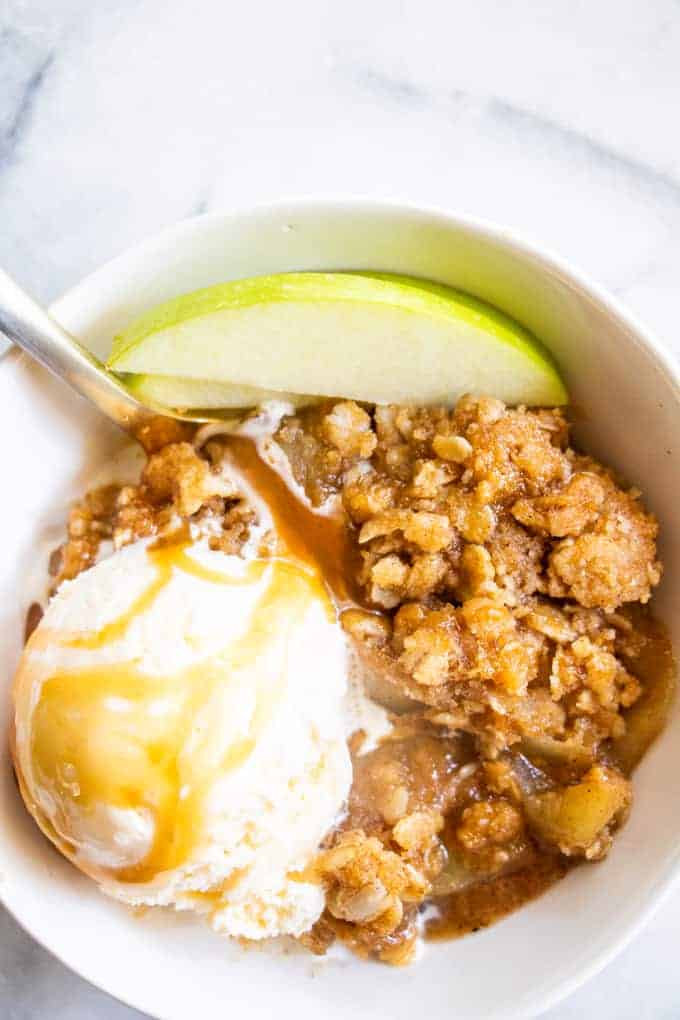 A bowl filled with caramel apple crisp, topped with ice cream drizzled with caramel and slices of apple. 