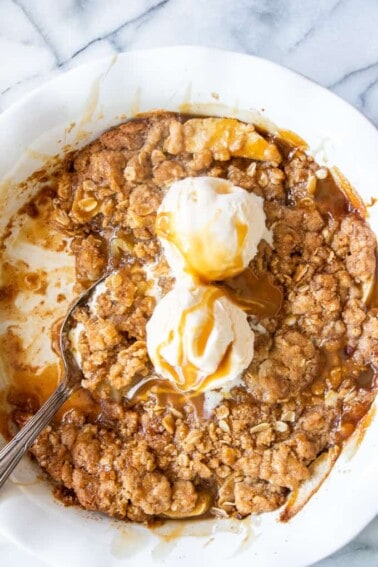 A pie dish filled with caramel apple crisp topped with melty ice cream and drizzled with more caramel sauce.
