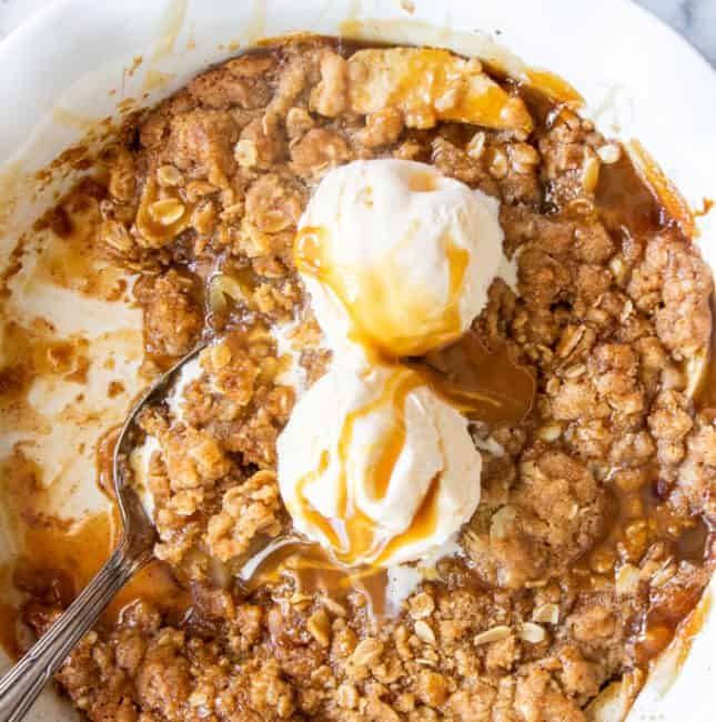 A pie dish filled with caramel apple crisp topped with melty ice cream and drizzled with more caramel sauce.