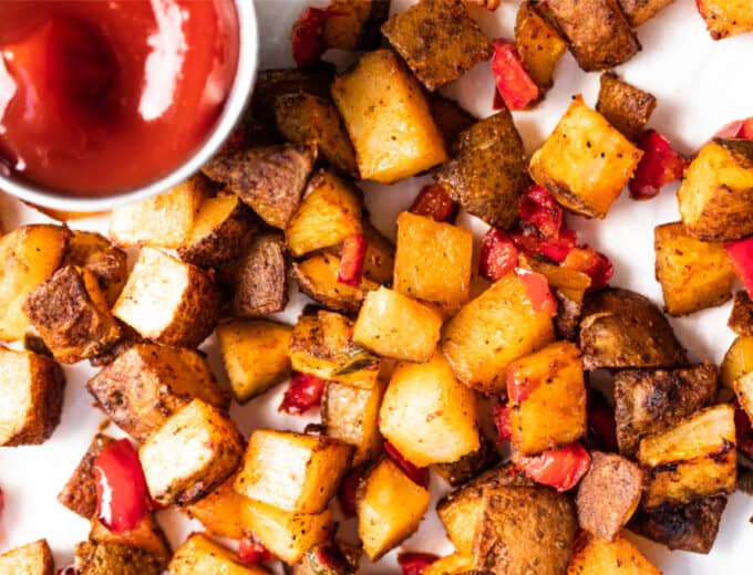 Baked, crispy breakfast potatoes mixed with red bell pepper and jalapeño spread onto a piece of parchment paper served with a cup of ketchup.