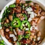 A bowl of charro beans topped with sliced jalapeno, diced onion and crispy bacon.