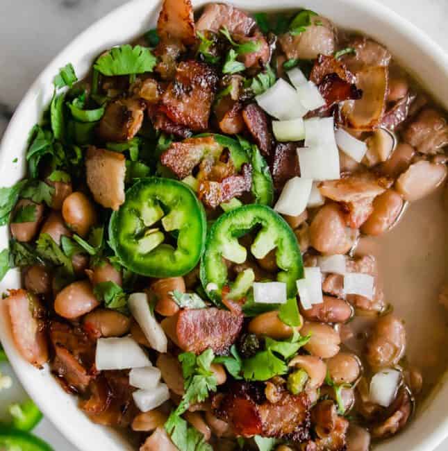 A bowl of charro beans topped with sliced jalapeno, diced onion and crispy bacon.