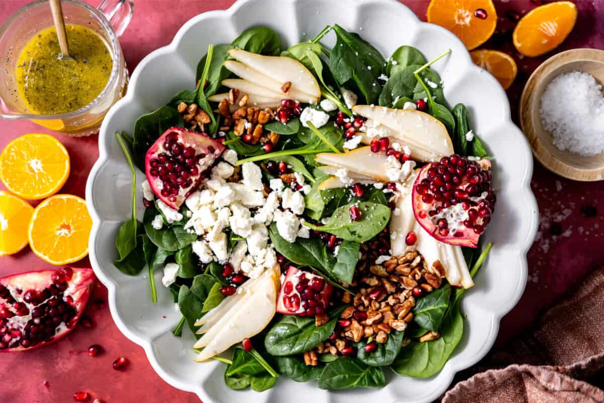 Ingredients needed for a Christmas salad in a white serving bowl with a jar of dressing on the side. 