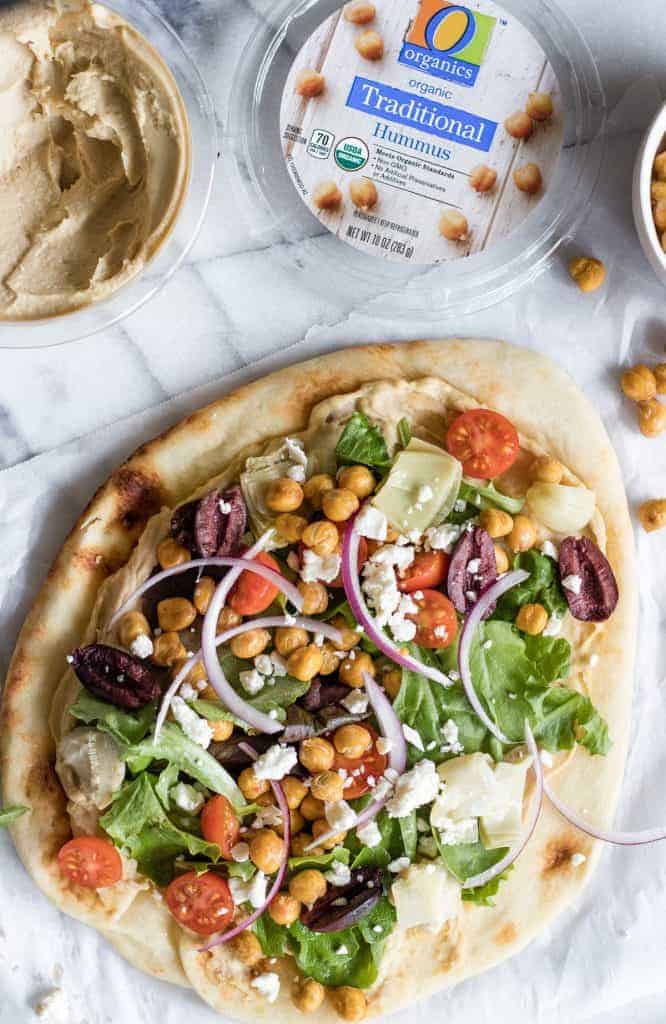 Completed flatbread pizza topped with hummus, salad, diced tomatoes, kalamata olives, red onion, roasted chick peas and artichokes.