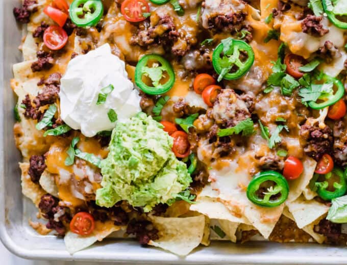 Sheet pan nachos layered with ground beef, cheese, tomatoes, cilantro, jalapeños, sour cream and guacamole.