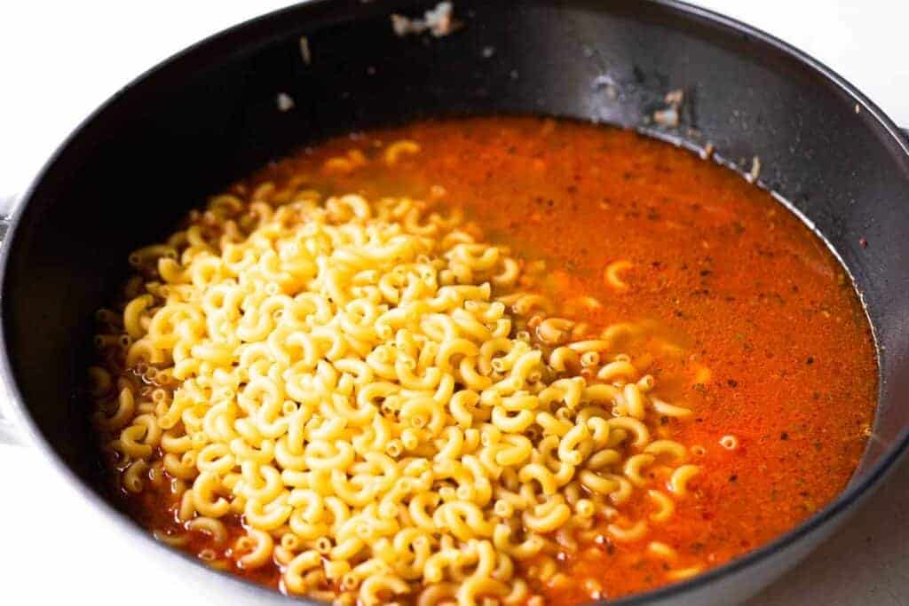 Macaroni noodles being added to red sauce in a skillet.