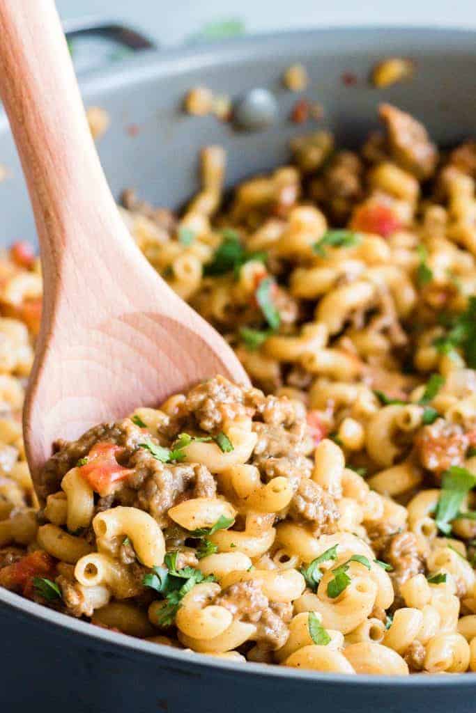 Wooden spoon scooping up cheesy one pot taco pasta, loaded with beef and tomatoes. Sprinkled with cilantro.