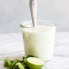 A jar full of avocado ranch dressing with a spoon dipped in. Surrounded by fresh herbs and half a lime.