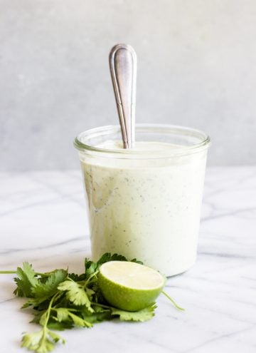 A jar full of avocado ranch dressing with a spoon dipped in. Surrounded by fresh herbs and half a lime.
