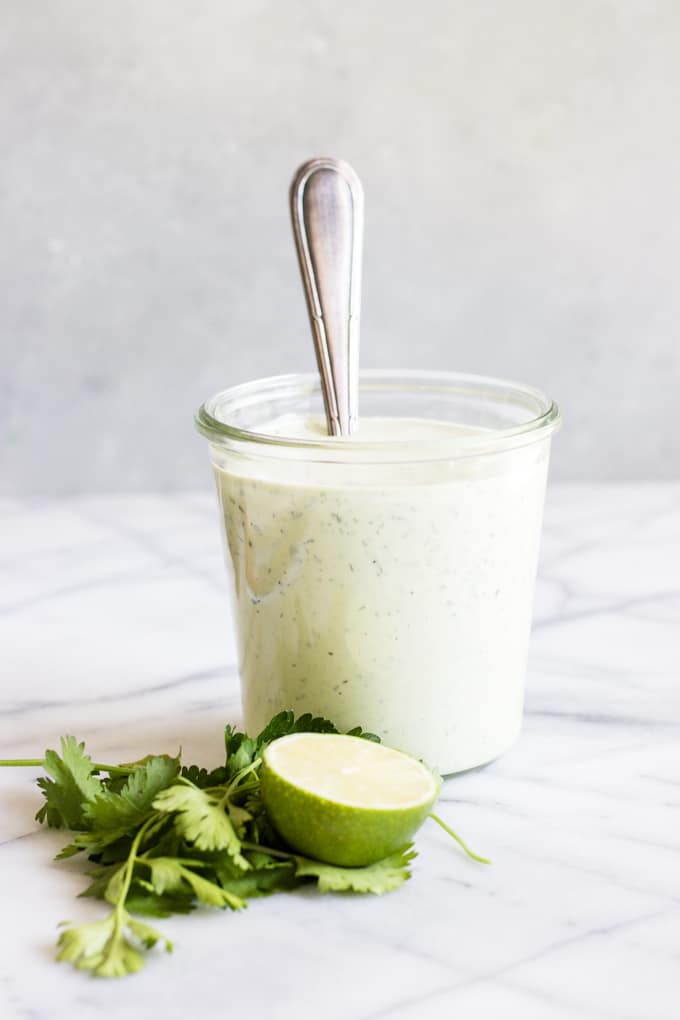 Een pot vol avocado ranch dressing met een lepel erin gedoopt. Omringd door verse kruiden en een halve limoen.