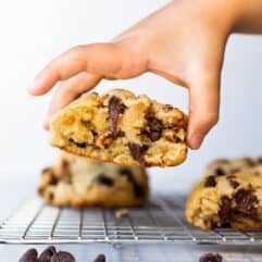 Super thick chocolate chip cookie being picked up by a hand off a cooling wire rack.