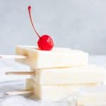 Stacked creamy coconut milk pina colada popsicles topped with a red cherry.