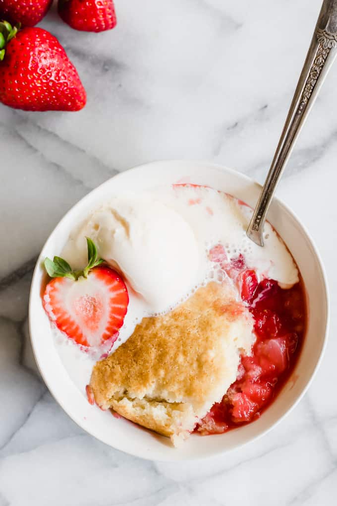 A bowl filled with strawberry cobbler, topped with ice cream and a half strawberry on top.