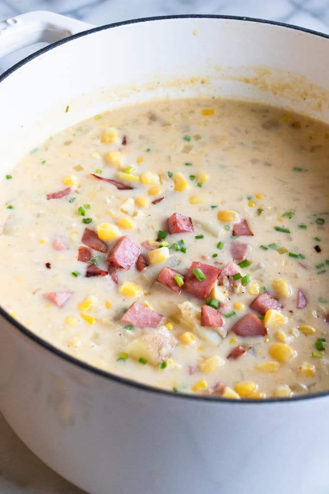 A white dutch oven filled with corn chowder, loaded up with bits of potato and ham.