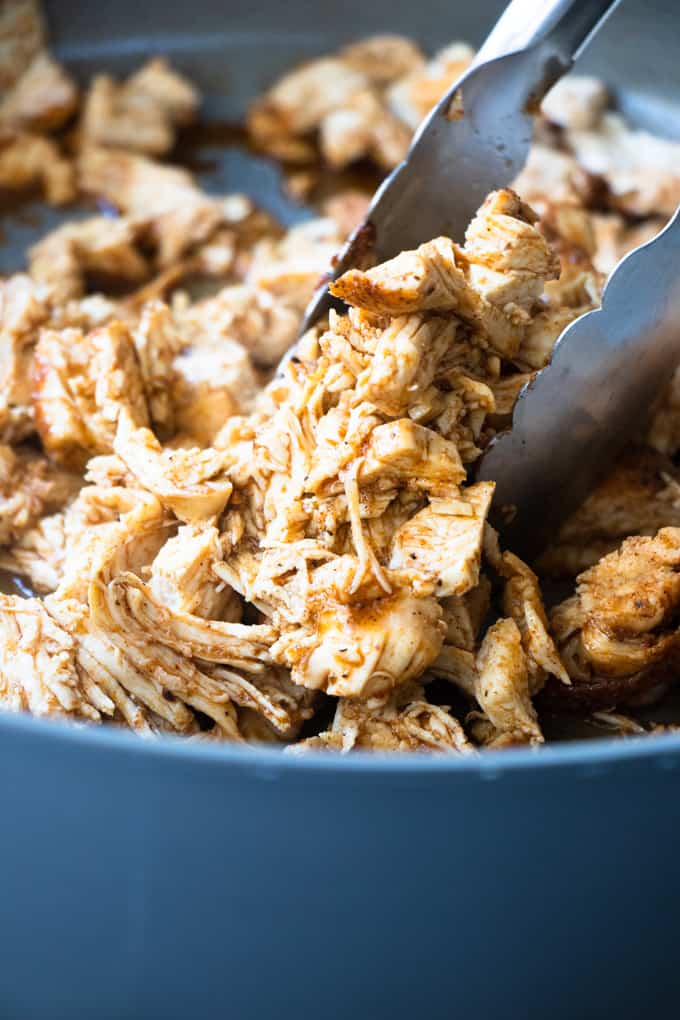 Tongs pulling up cooked, shredded chicken to pile up into tortillas for chicken tacos.