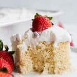A slice of tres leches cake, topped with whipped cream and a strawberry with a bite missing.