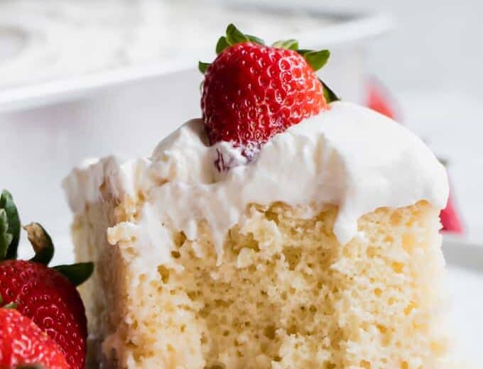 A slice of tres leches cake, topped with whipped cream and a strawberry with a bite missing.