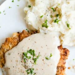 A plate with chicken fried steak and mashed potatoes, drizzled with cream gravy and garnished with cracked pepper and parsley.