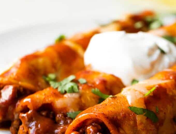 Homemade Beef Enchiladas served on a plate with diced cilantro and a swirl of sour cream on top.
