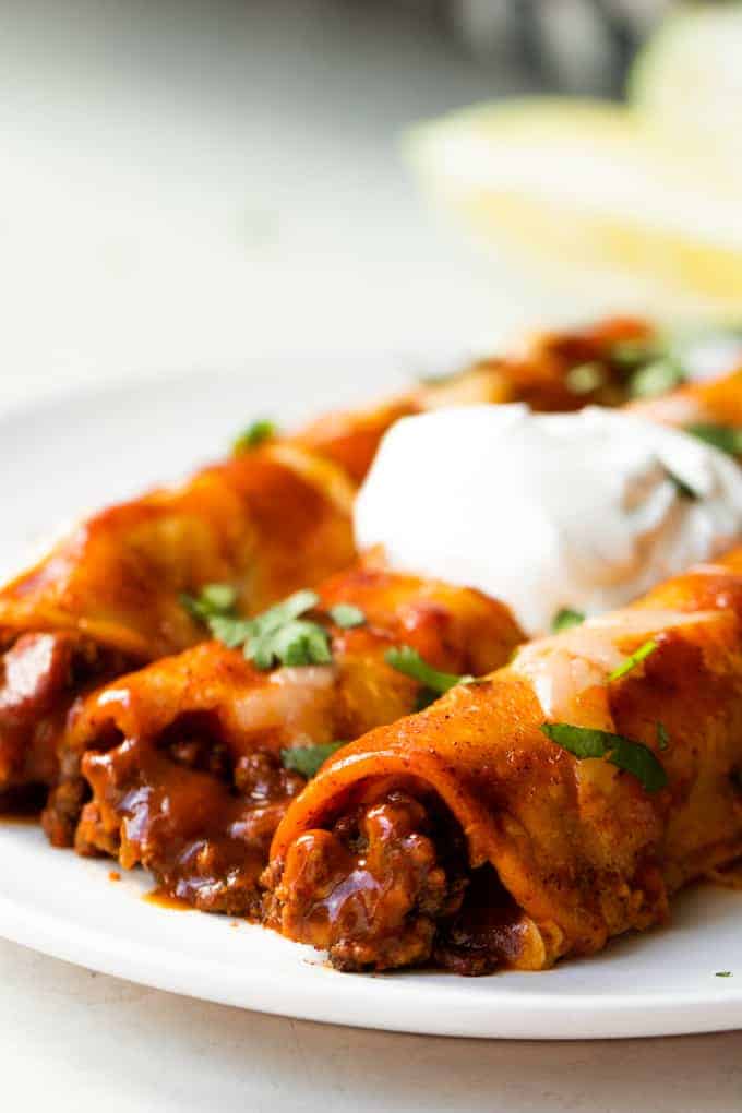 Homemade Beef Enchiladas served on a plate with diced cilantro and a swirl of sour cream on top. 