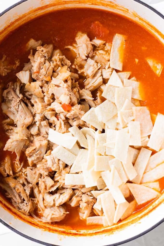 Pot of tortilla soup broth with shredded chicken and corn tortillas being added.