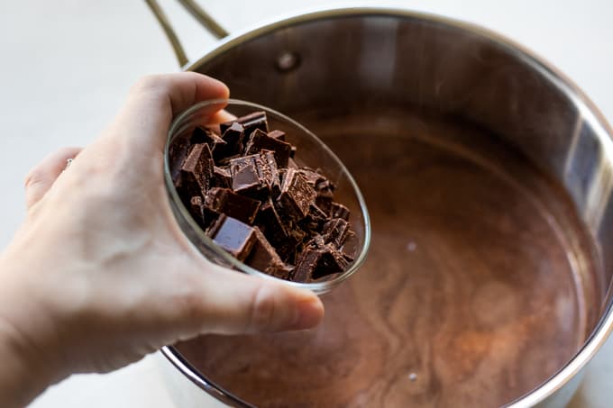 Hand pouring bowl of chopped up chocolate into a saucepan filled with hot chocolate. 
