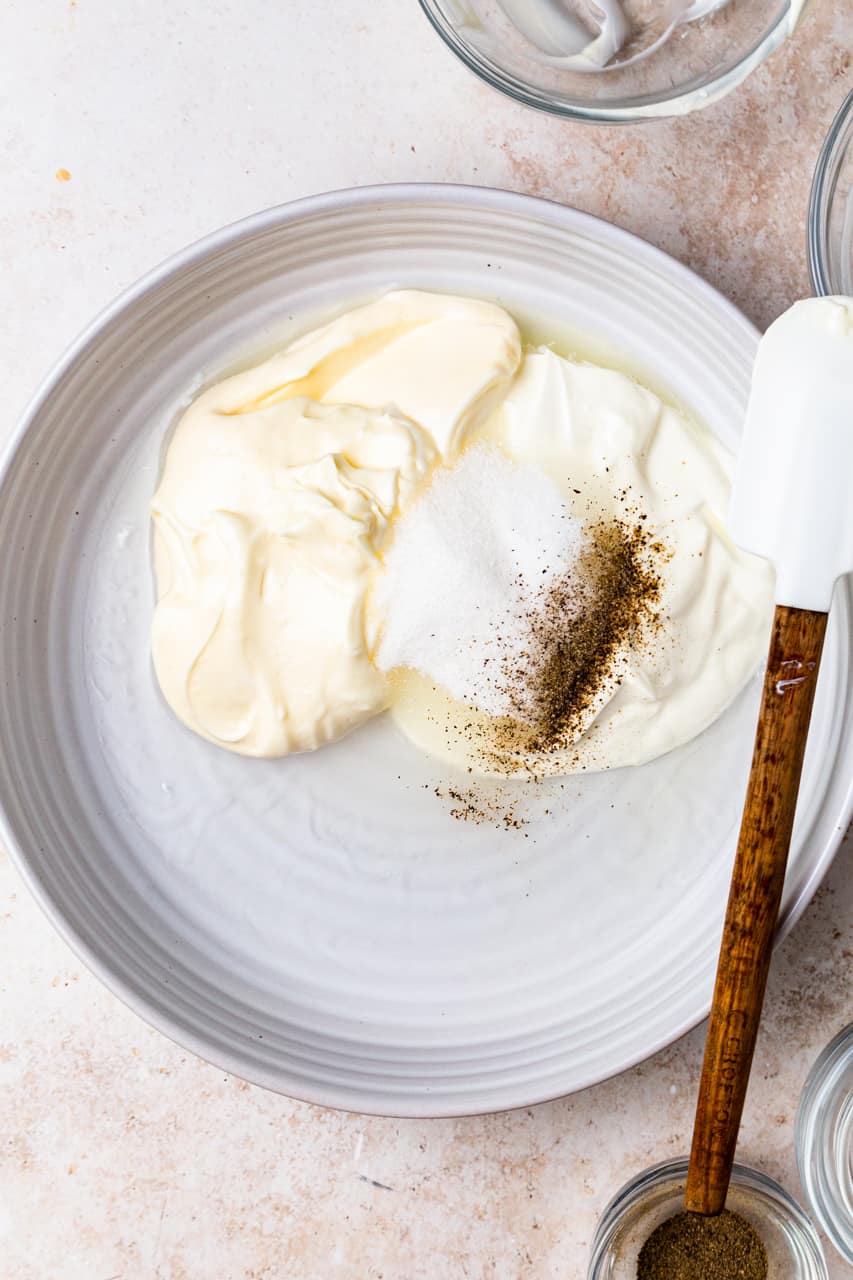 Mayonnaise, sour cream, vinegar and seasonings in a large white bowl with a white silicone spatula. 