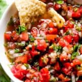 White bowl filled with pico de Gallo and a tortilla chip dipped in showing the juices.