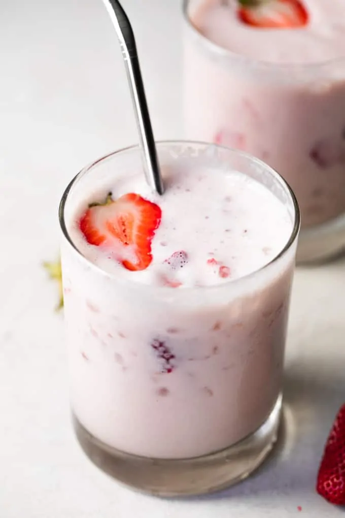 A glass filled with strawberry milk, garnished with a halved strawberry and served with a stainless steel straw.