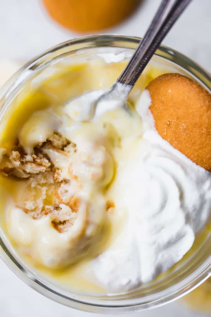 Overhead view of banana pudding in a glass showing vanilla pudding, topped with soft whipped cream, garnished with a Nilla wafer, a spoon digging in showing crumbled cookies in the pudding.
