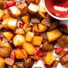 Baked, crispy breakfast potatoes mixed with red bell pepper and jalapeño spread onto a piece of parchment paper served with a cup of ketchup.