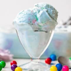 Glass ice cream cup filled with light blue bubble gum ice cream, container of ice cream in the background, and extra gumballs around the ice cream cup.