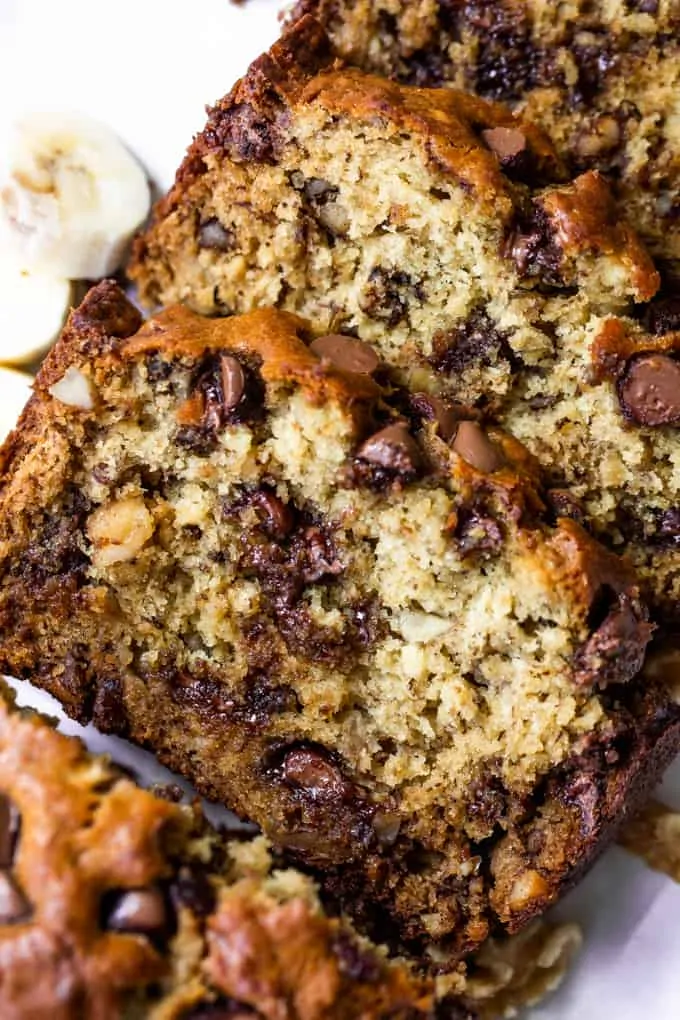 Sliced loaf of chunky monkey banana bread showing banana bread with chunks of walnut, and melted chocolate chips. Slices of bananas and walnuts on the side.