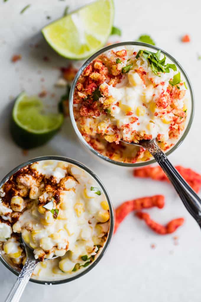 Mexican street corn in cups served with crunched cheetos and chili sauce, with lime wedges and diced cilantro on the side.