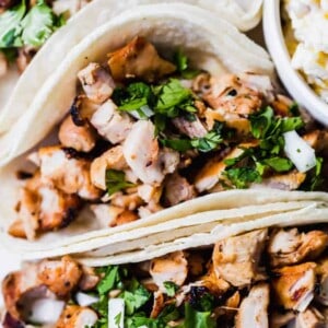 Up close image of diced up grilled chicken in soft white corn tortillas topped with diced white onion and cilantro. A bowl of street corn on the side.