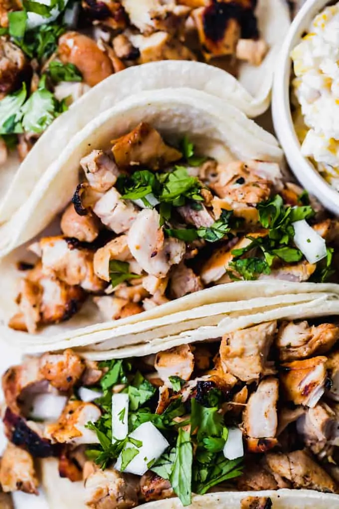 Up close image of diced up grilled chicken in soft white corn tortillas topped with diced white onion and cilantro. A bowl of street corn on the side. 