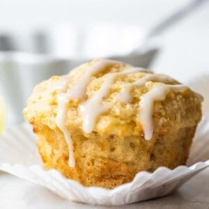 Lemon muffin sitting in a white cupcake liner, with a light white drizzle on top. Bowl of icing with a spoon behind the muffin.