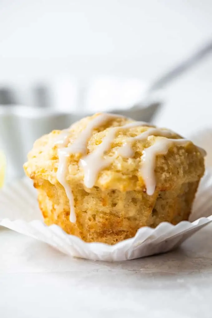 Lemon muffin sitting in a white cupcake liner, with a light white drizzle on top. Bowl of icing with a spoon behind the muffin.