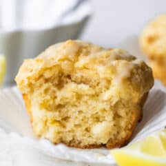 Horizontal image of baked lemon muffin with a bite missing showing fluffy texture. Lemon wedges on the side and a bowl of icing behind the muffin.