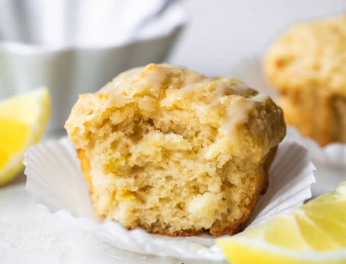 Horizontal image of baked lemon muffin with a bite missing showing fluffy texture. Lemon wedges on the side and a bowl of icing behind the muffin.