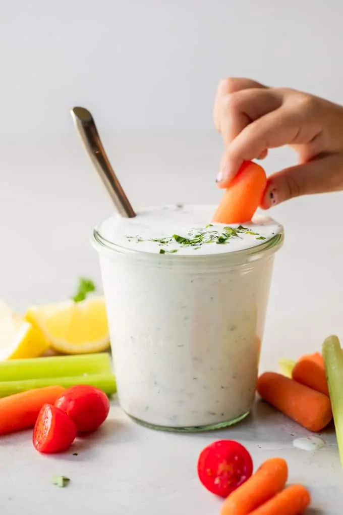Small hand with chipped nail polish dipping a baby carrot into a jar filled with homemade ranch dressing. 