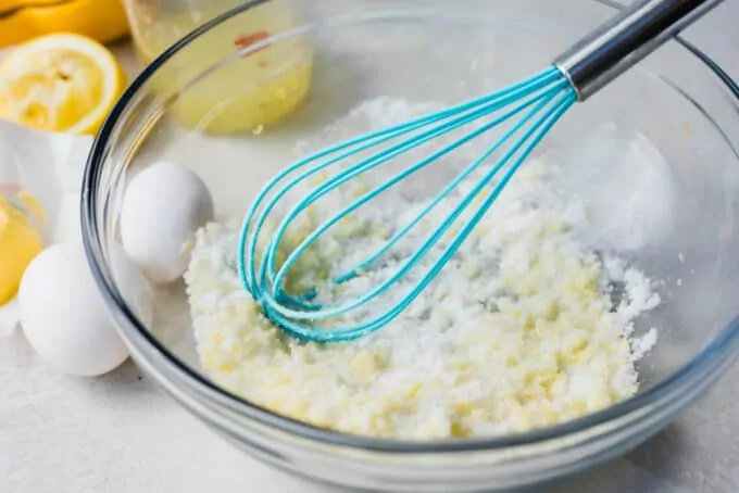 Glass bowl filled with sugar and lemon zest with eggs on the side and a blue whisk.