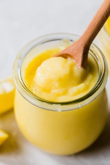 Tulip glass jar filled with bright yellow homemade lemon curd with a wooden spoon scooping it out.